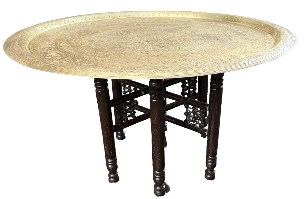 A round brass tray rests on a dark wooden table with four legs, set on a gravel surface next to a house with beige siding.
