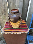 A rustic brown ceramic pot sits on a colorful striped textile with lace trim, adorned with wooden beads and leather straps against a wooden fence.