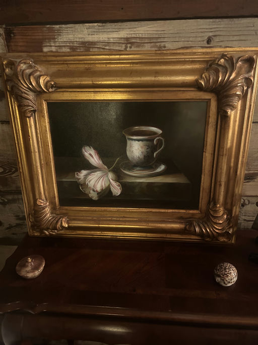 A gold-framed painting depicts a delicate teacup with coffee, resting on a wooden surface beside a pink and white flower. The background is rustic.