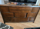 A wooden dresser with a modern design features a geometric pattern on the front. The dresser sits on a wooden floor next to a TV and a hat.