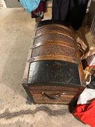 A vintage trunk with a curved lid, featuring intricate metal accents and a wooden base, sits on a concrete surface surrounded by clutter.