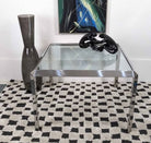 A glass-top coffee table with metal legs sits on a checkered rug. A tall gray vase and a black sculpture are displayed on the table beside a colorful painting.