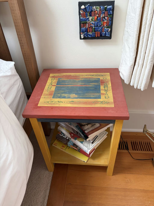 A colorful, hand-painted side table with a blue and red top holds a stack of various books on its lower shelf. A mosaic artwork hangs nearby.