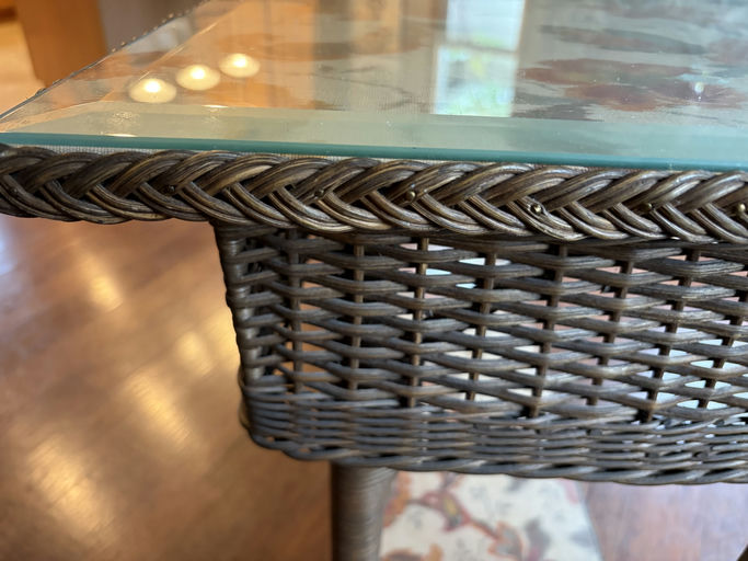 A close-up of a wicker table's edge with a glass top, showcasing intricate braided patterns and a wooden floor in the background.