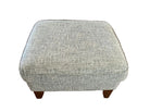 A square upholstered ottoman with a textured gray fabric sits on a hardwood floor. It has wooden legs and is located in a cozy living space.