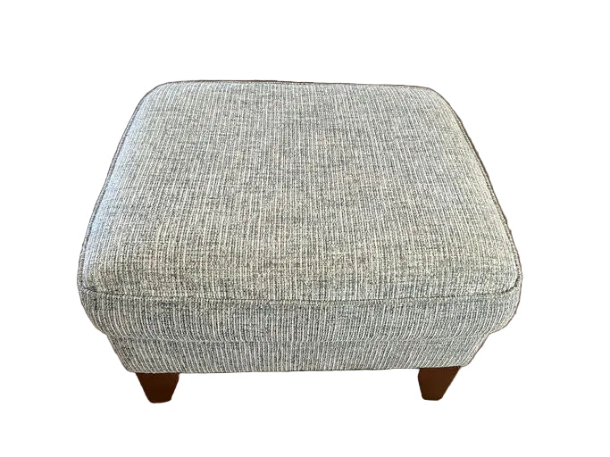 A square upholstered ottoman with a textured gray fabric sits on a hardwood floor. It has wooden legs and is located in a cozy living space.