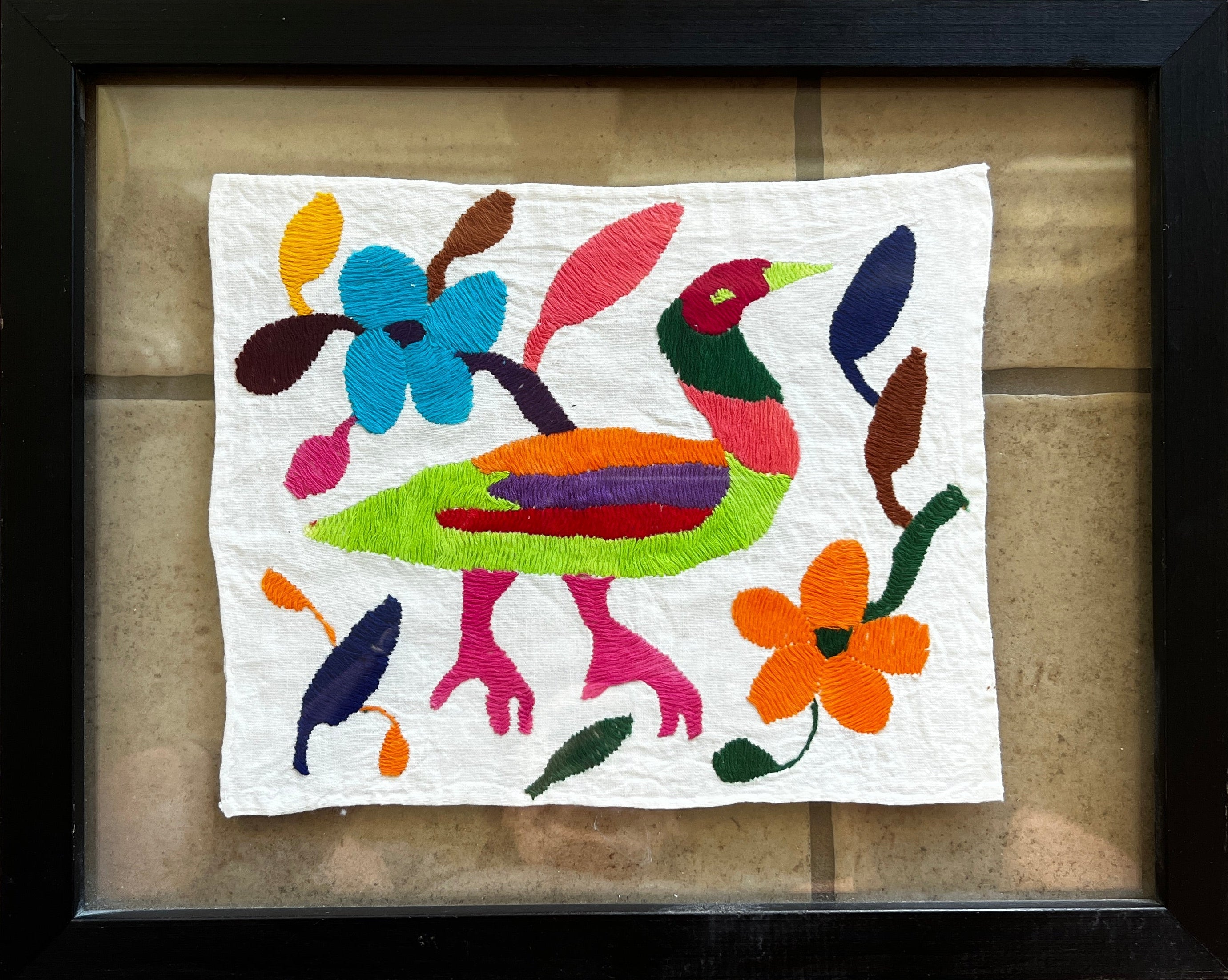A colorful embroidered artwork featuring a stylized bird surrounded by flowers and vibrant shapes, framed against a tiled background.