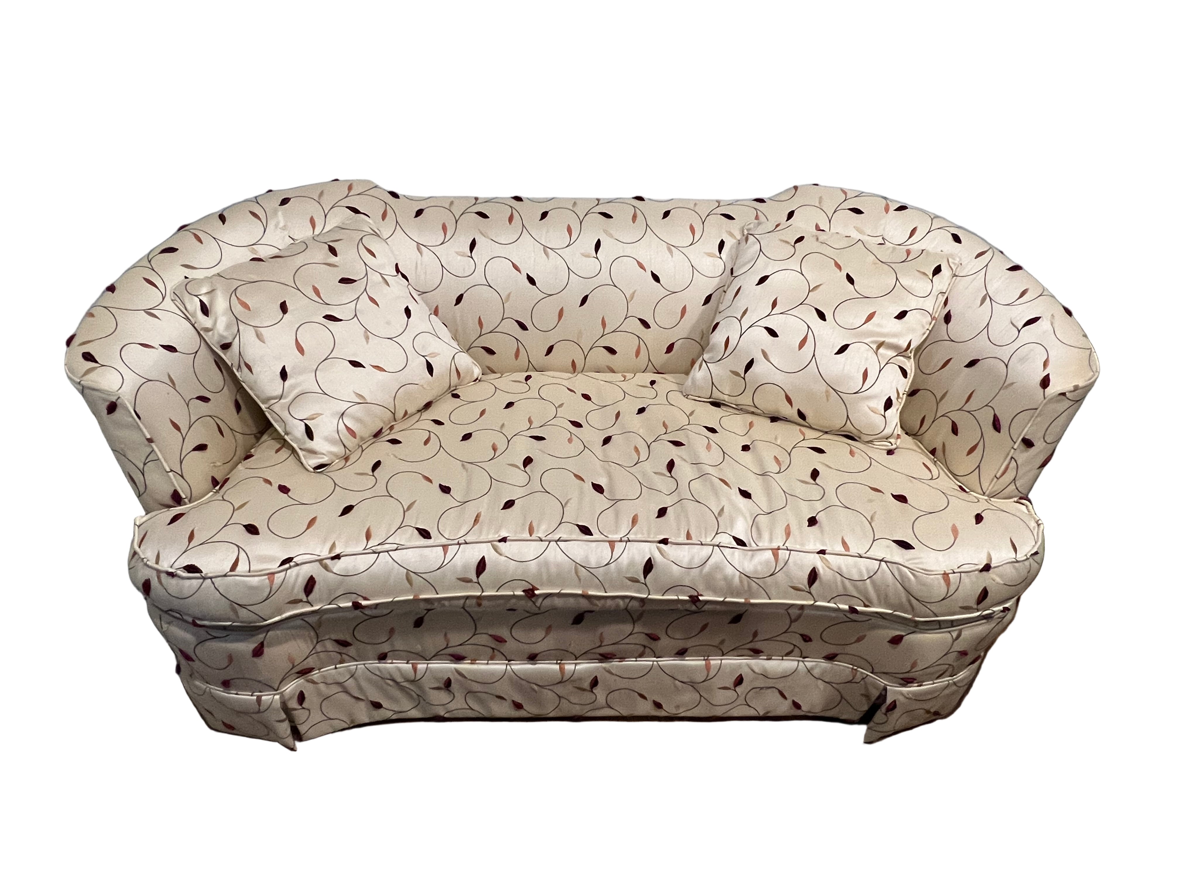 A plush, two-seater sofa with cream fabric featuring a subtle pattern of maroon leaves, accompanied by two matching, square cushions.