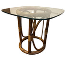 A small, round table with a glass top and a woven rattan base, set on a wooden floor with a wall behind it. Simple, modern design.