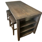 A rustic wooden table with two upholstered stools is positioned in a dining area, featuring open shelves underneath and a warm wooden floor.