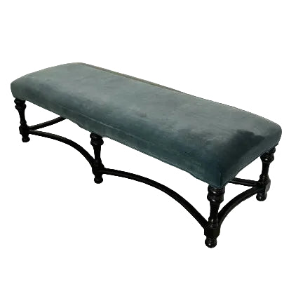 A long, upholstered bench with a textured teal fabric rests against a white wall, set on a carpeted floor. The black legs add elegance.