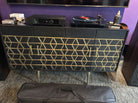 A stylish black cabinet with gold geometric patterns houses a turntable and audio equipment, accompanied by a black bag on the floor.