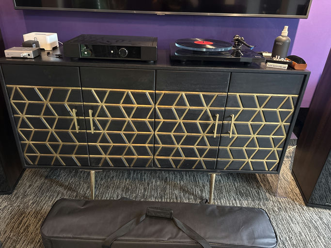 A stylish black cabinet with gold geometric patterns houses a turntable and audio equipment, accompanied by a black bag on the floor.