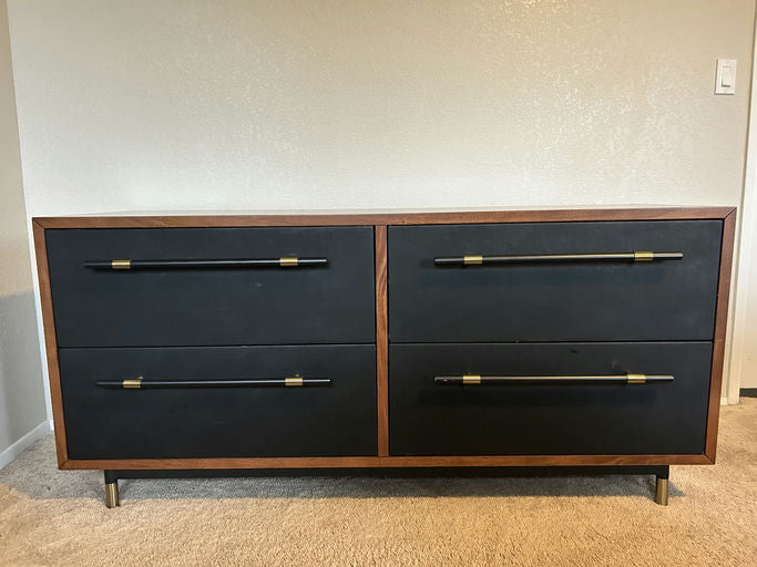 A modern dresser with a wooden frame, featuring four black drawers, each with sleek black handles accented by gold hardware. Set on metal legs.