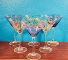 A collection of colorful, patterned martini glasses arranged in a semi-circle against a vibrant turquoise background, resting on a wooden surface.