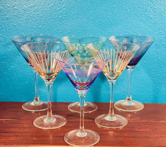 A collection of colorful, patterned martini glasses arranged in a semi-circle against a vibrant turquoise background, resting on a wooden surface.