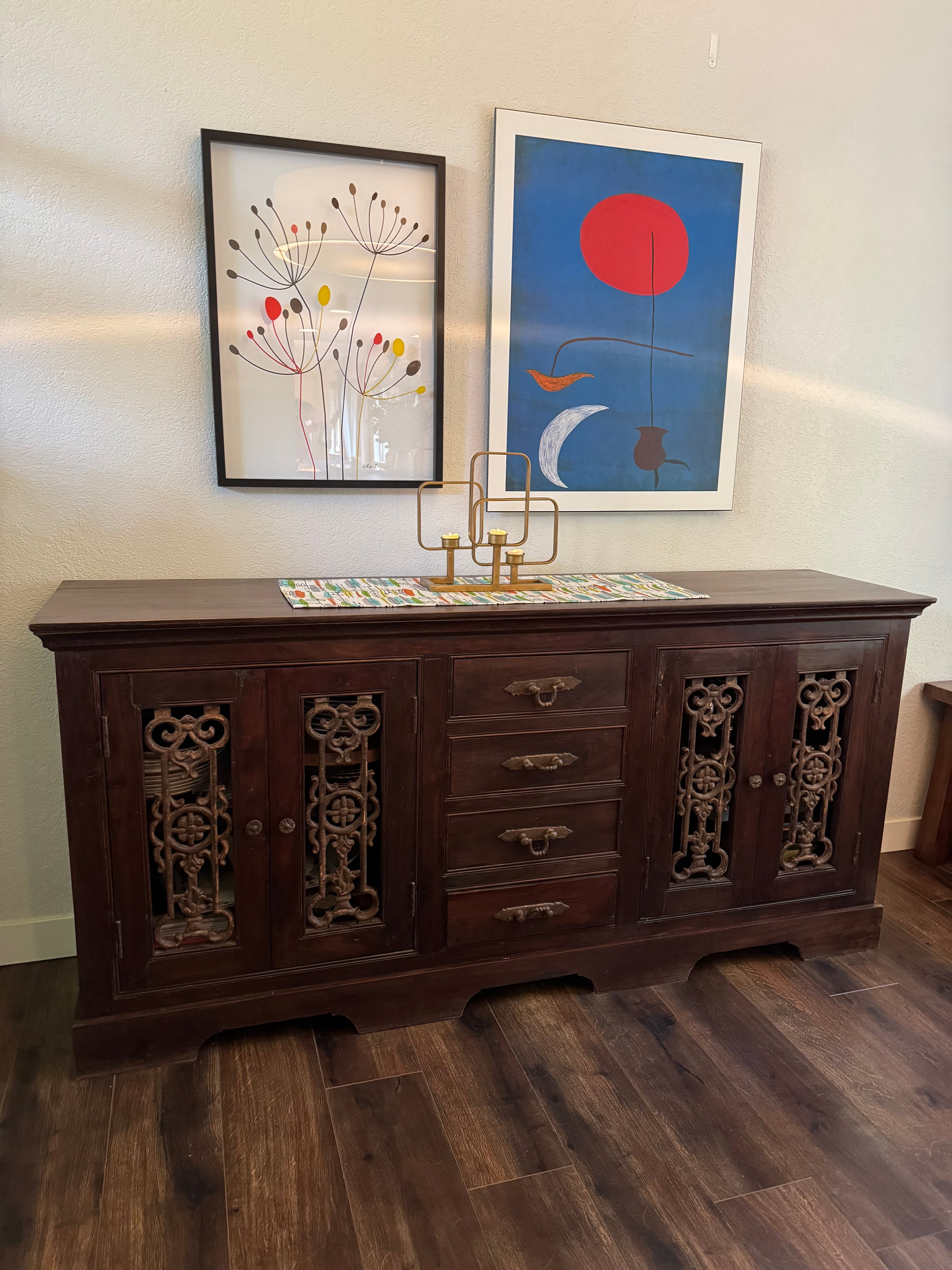 A wooden sideboard with intricate carvings, topped with a colorful table runner. Two framed artworks hang above it: one abstract, one floral.