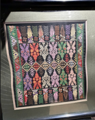 Colorful textile artwork features intricate geometric patterns and stylized motifs in vibrant hues, framed on a black background.