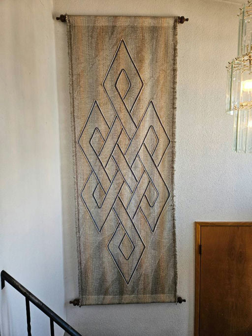 A woven wall hanging featuring an intricate geometric pattern in earthy tones, displayed against a light wall near a staircase and wooden door.