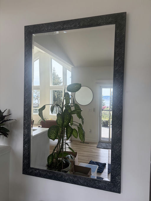 A decorative mirror reflects a bright room with large windows, a potted plant, and a circular mirror. Natural light fills the space.