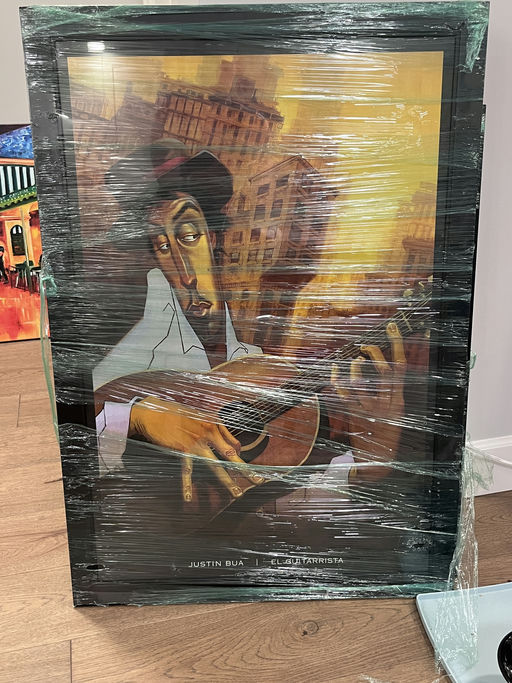 A framed illustration by Justin Bua titled "El Guitarrista," featuring a guitarist surrounded by an urban backdrop, wrapped in plastic.