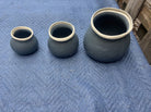 Three ceramic pots of varying sizes sit on a blue quilted surface. The pots are dark with cream-colored rims, showcasing smooth, rounded shapes.