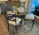 A round glass dining table with four ornate black metal chairs, set on a patterned rug. A small potted plant sits in the center, adding a touch of decor.