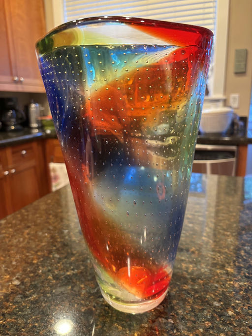 A colorful glass vase with swirling red, blue, green, and yellow hues, decorated with small bubbles, sitting on a dark granite counter.