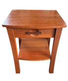 A wooden side table with a single drawer and open shelf sits on a concrete floor, surrounded by cardboard boxes and a green blanket.