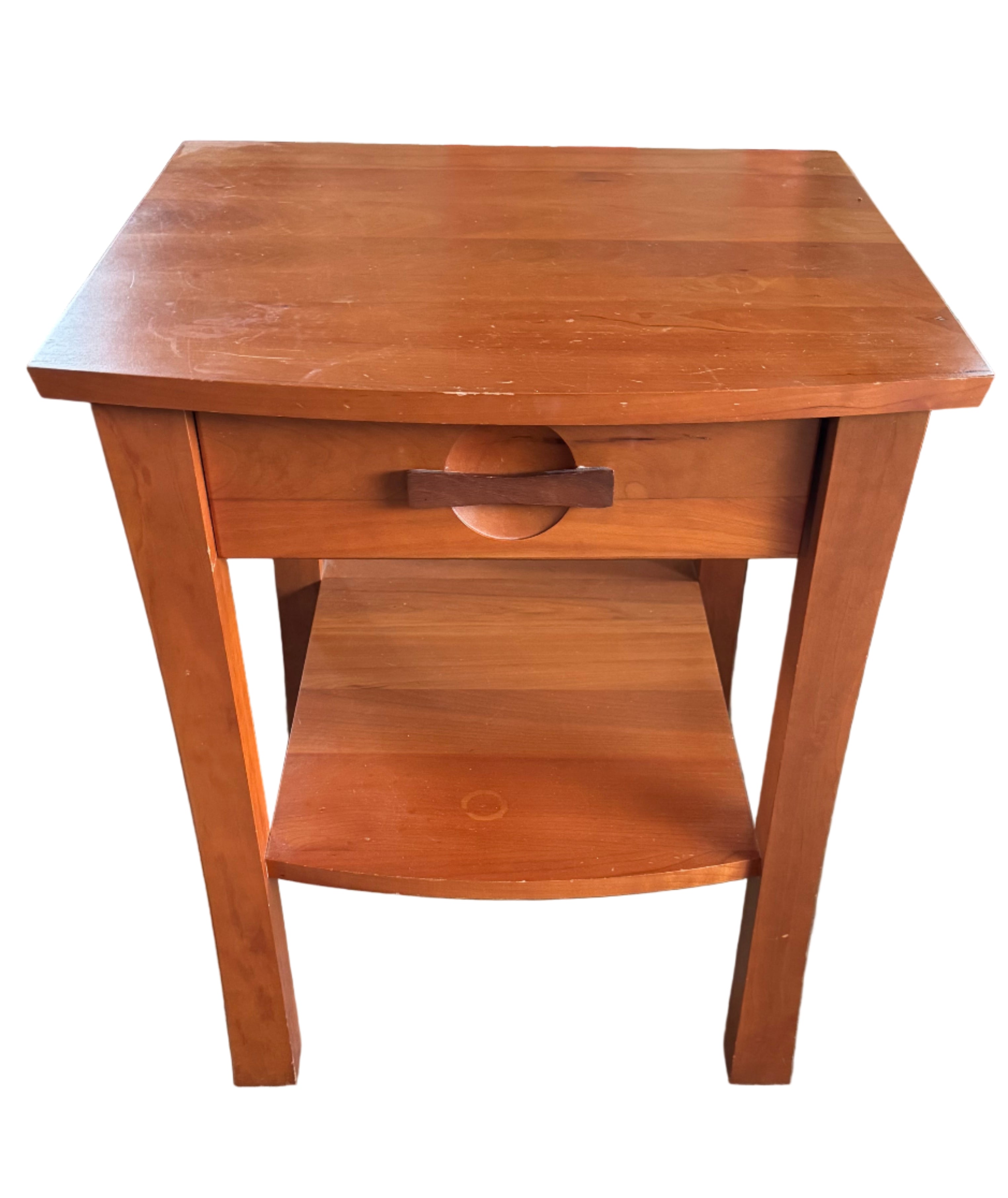A wooden side table with a single drawer and open shelf sits on a concrete floor, surrounded by cardboard boxes and a green blanket.
