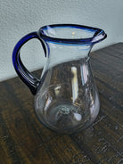A clear glass pitcher with a blue rim and handle rests on a wooden table, showcasing its elegant design against a textured background.