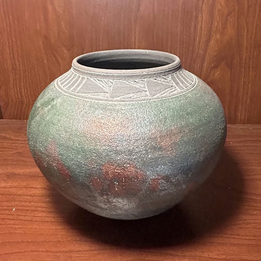 A round, textured pot with a dark green and red patina, featuring a decorative band around the rim, sits on a wooden surface.