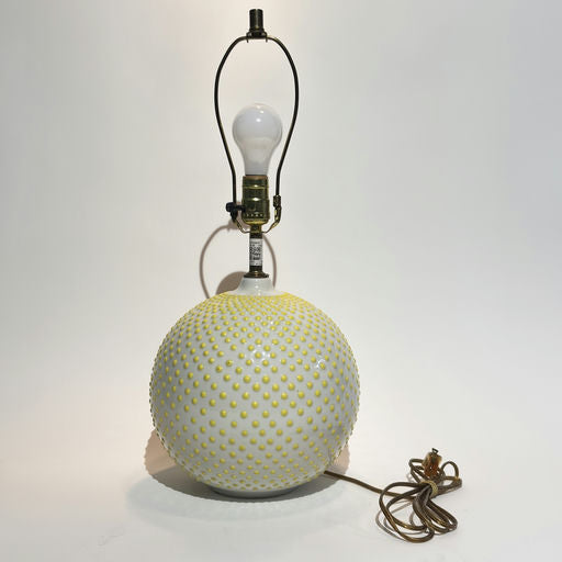 A lamp with a spherical, white base covered in yellow dots, featuring a light bulb socket and a brown electrical cord, set against a neutral background.