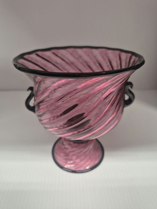 A decorative glass vase with a twisting pink design and black accents, featuring two handles and a flared rim, resting on a round pedestal base.