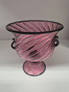 A decorative glass vase with a twisting pink design and black accents, featuring two handles and a flared rim, resting on a round pedestal base.