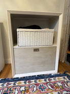A white shelving unit with a woven storage basket on top and a wooden drawer below, placed on a colorful patterned rug. Soft lighting and decor visible.