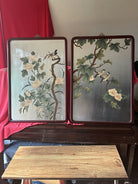 Two decorative panels featuring stylized floral designs and perched birds, set against a neutral background, displayed on a wooden table.