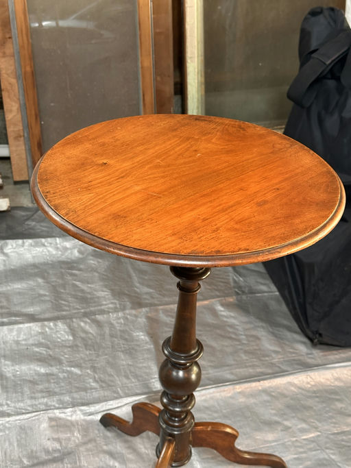 A wooden side table with a round top and a decorative, turned pedestal base, set against a backdrop of blurred furniture and a dark cloth.