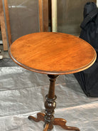 A wooden side table with a round top and a decorative, turned pedestal base, set against a backdrop of blurred furniture and a dark cloth.