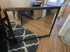A wooden-framed mirror leans against a cabinet on a hardwood floor, reflecting part of a room with a rug, chair, and various household items.