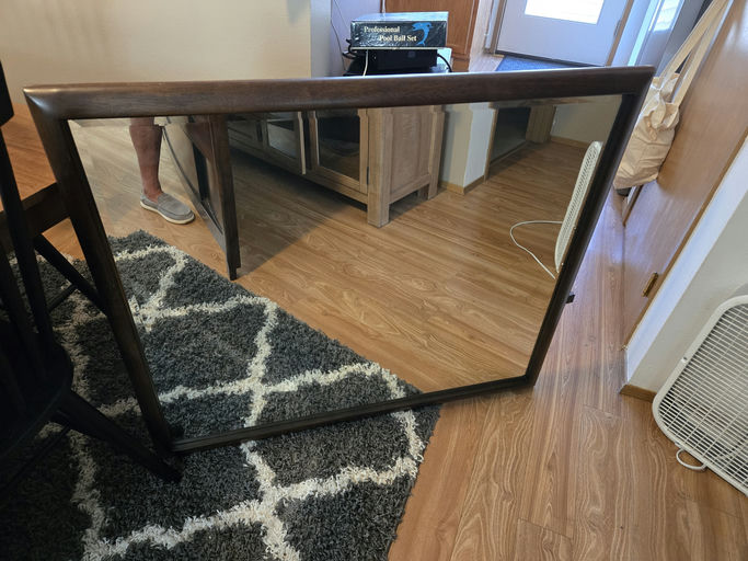 A wooden-framed mirror leans against a cabinet on a hardwood floor, reflecting part of a room with a rug, chair, and various household items.