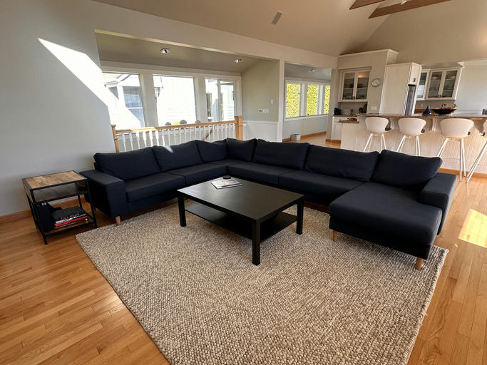 A cozy living room with a dark sectional sofa, black coffee table, and light wooden flooring. Sunlight streams in through large windows.