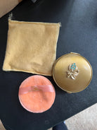 A gold compact mirror with a floral design, an orange puff, and a yellow cloth are arranged on a dark surface. The cloth shows stitching details.