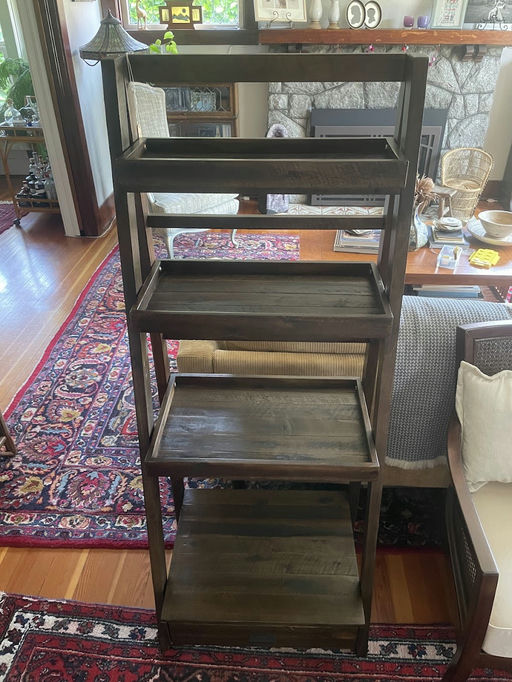 Wooden shelving unit with five open shelves in a cozy living room setting, featuring a patterned rug, a stone fireplace, and surrounding furniture.