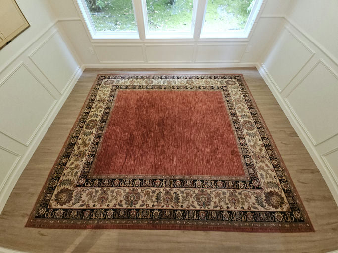 A large red area rug with intricate patterned borders sits on a wooden floor, surrounded by light wooden paneling and large windows.