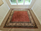 A large red area rug with intricate patterned borders sits on a wooden floor, surrounded by light wooden paneling and large windows.