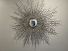 A decorative sunburst mirror with a silver frame and protruding branches, reflecting a person standing in front of it against a light wall.
