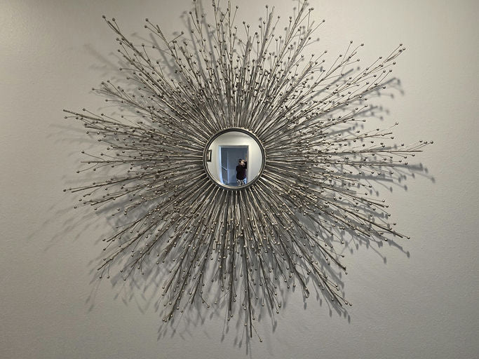 A decorative sunburst mirror with a silver frame and protruding branches, reflecting a person standing in front of it against a light wall.