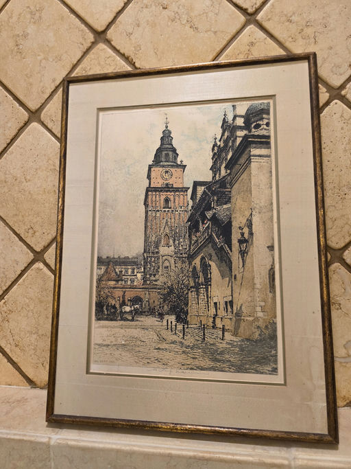 A framed vintage artwork depicting a tall clock tower and buildings along a cobblestone street, set against a textured tile background.