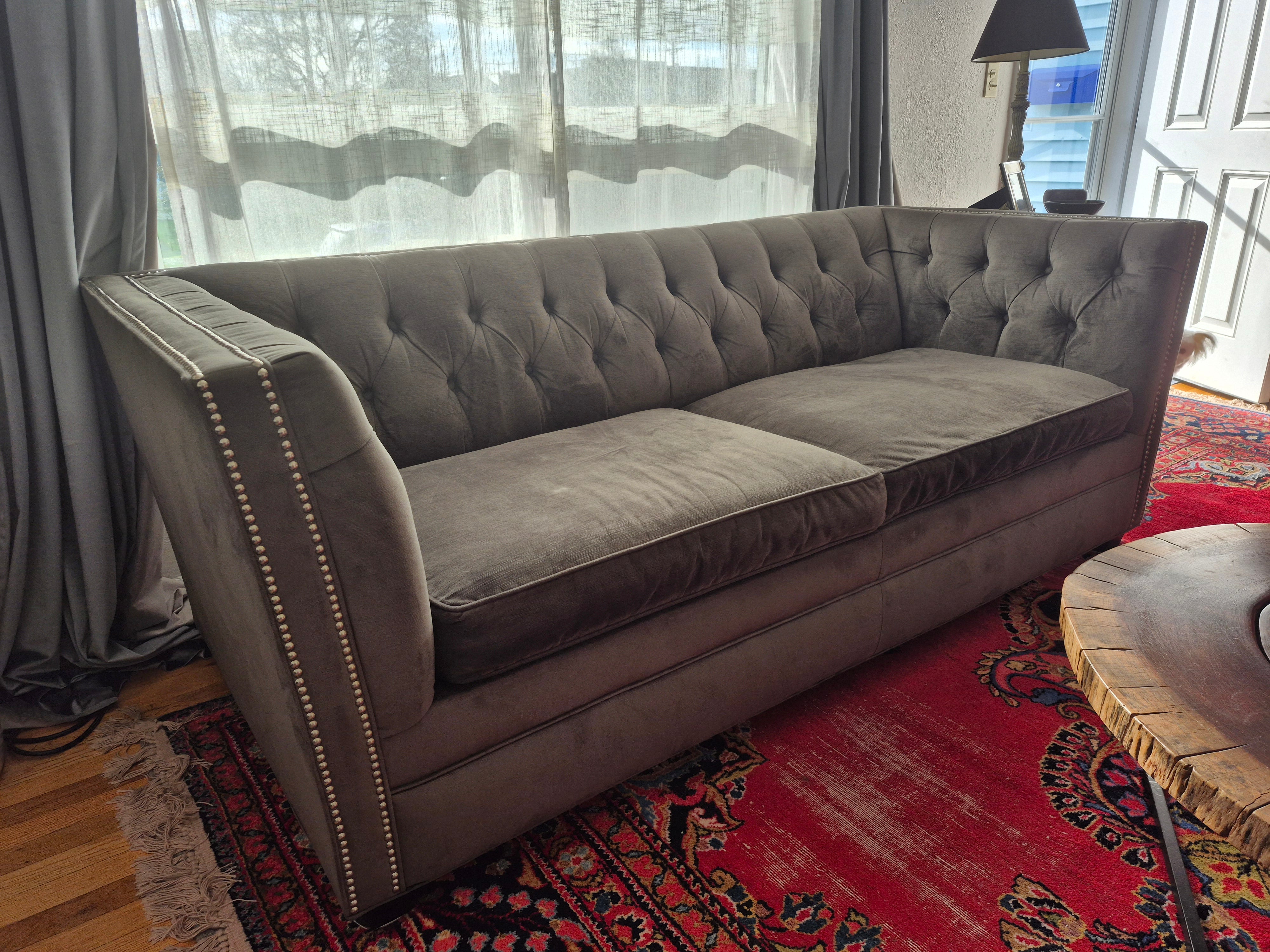 A stylish, tufted gray sofa with decorative nailhead trim sits on a colorful patterned rug, near sheer curtains and a doorway. Natural light pours in.